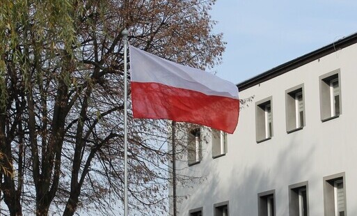 Flaga RP na tle błękitnego nieba, drzewa i budynku